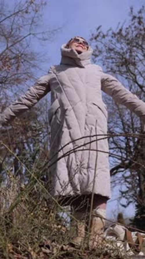Une femme âgée verticale et détendue dans une veste aux bras écartés regarde le soleil dans le parc