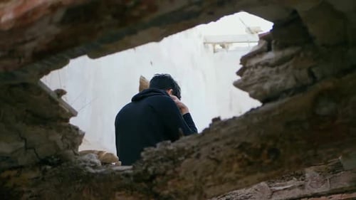 Young man, teenager, sitting high on a beam in an abandoned house puts on his hood. Young sad, depre