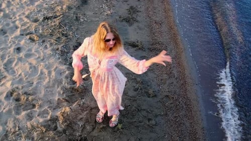 Top view of young woman dancing on sand near the seashore with a long dress flowing in the wind
