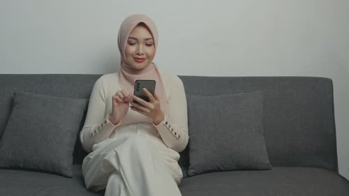 Pretty young Asian Muslim woman in hijab using smartphone while sitting down on a sofa.