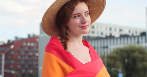 Retrato de uma bela jovem com um chapéu com longos cabelos ruivos cobrindo-se na bandeira LGBT enquanto