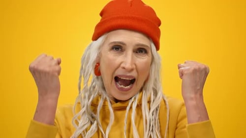 Cheerful senior woman raising fists in celebration on yellow studio background