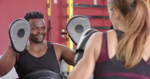 A fit African American man is training a young Caucasian woman in boxing at the gym