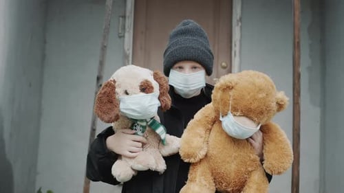 A Sad Child Stands on the Threshold of the House Holding Two Toys in Protective Masks