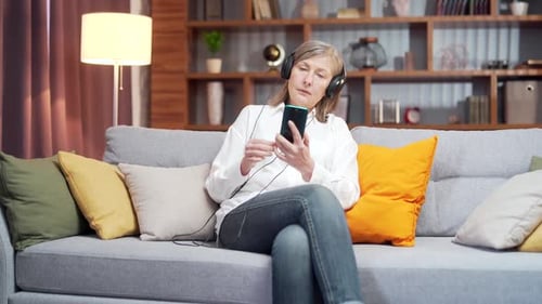 Senior woman listening music on the couch at home. Happy mature old female wear headphone holding