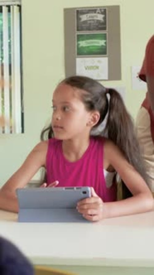 Vertical video: Leaning teacher checking girl in pink top resuming tapping gray tablet in classroom