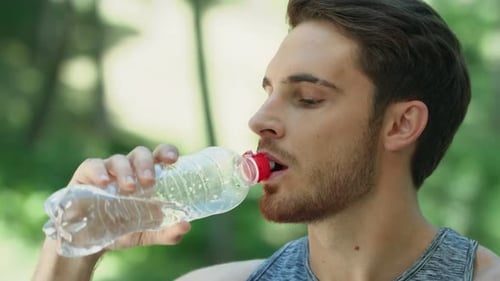 Thirsty sport man drinking refreshing water from bottle after workout in summer park