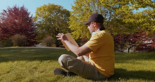 Mann liest ein Buch und sitzt bei Sonnenuntergang im Park im Gras