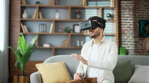 Young adult man works using VR glasses in virtual reality simulator sitting on sofa in living room