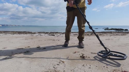Man Using Metal Detector on Sandy Beach Near the Sea Closeup View of Legs and Device Under Bright