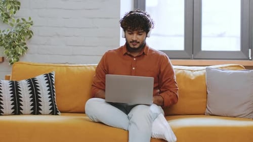 Young Man Working on Laptop While Sitting Comfortably on a Bright Yellow Sofa in a Cozy Indoor Space