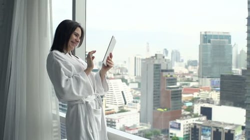 Smiling young woman in bathrobe uses tablet by window overlooking the city