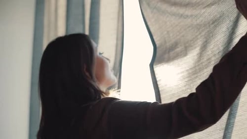 Woman Opens Curtains Letting Sunlight into Room