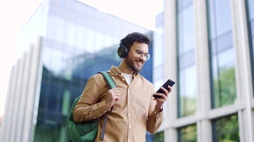 Handsome smiling student with a backpack uses a smartphone while walking in the campus space near