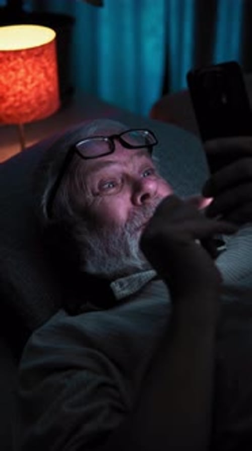 In Dark Old Man with Glasses Lies in Bed and Surfing News on His Phone