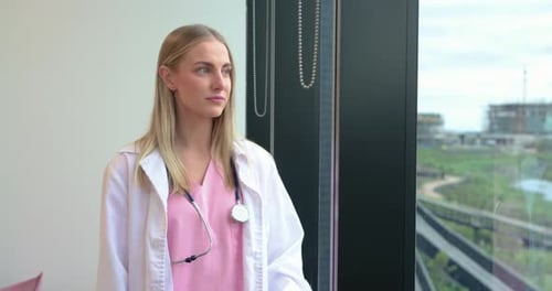 Erwachsene Ärztin in rosa Peelings mit Stethoskop steht am Klinikfenster und schaut sich um und lächelt