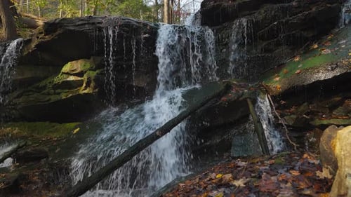 Water cascades over mossy rocks in a serene forest scene with colorful autumn leaves