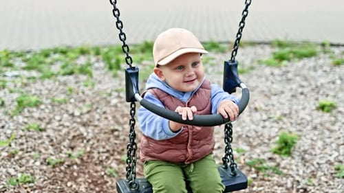 Happy toddler swings on a swing outside. A boy of 2 years old in a cap and a warm vest swings