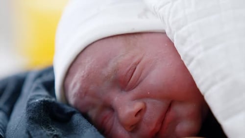 Face of a newborn right after birth. Tiny baby in white cap wants to cry and blinking eyes. Close up