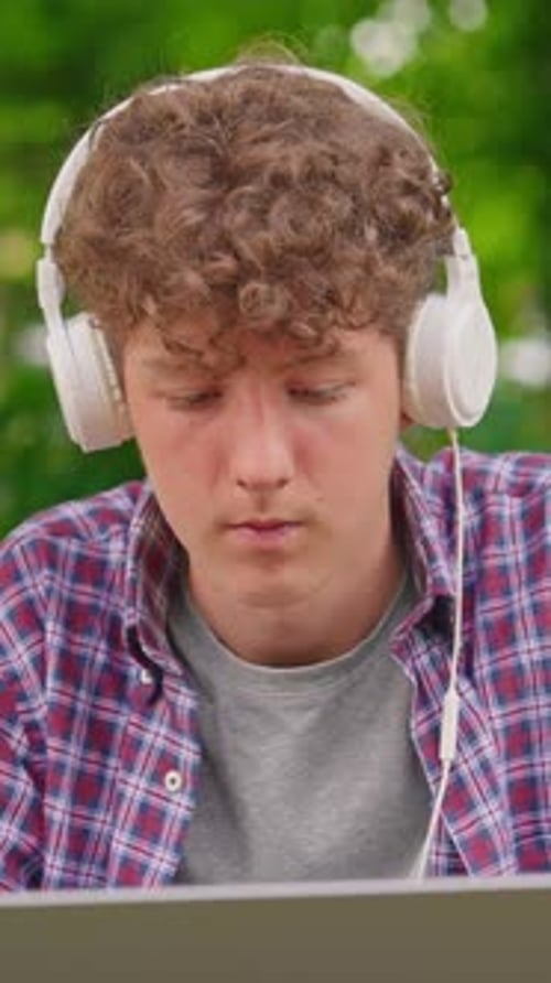 Vertical Video Curly Teenage Boy Listening to Music on Headphones and Works on a Laptop While