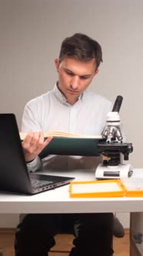 Vertical Video Student Focusing on a Textbook While Sitting at a Desk with a Microscope and Laptop