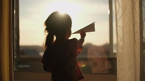 A Child Plays with a Paper Plane Near the Window of the House During Sunset