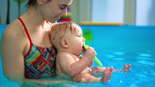 Woman is holding a little cute baby boy in the swimming pool. Lovely kid is chewing the toy.