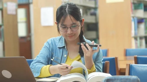 Asiatischer Student, der in der Bibliothek mit Laptopbüchern recherchiert, ein Projekt erstellt und sich auf die Prüfung vorbereitet.