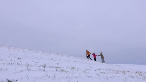 A Young Family of Three is Walking on a Snowy Hill in a Field Against a Gray Sky