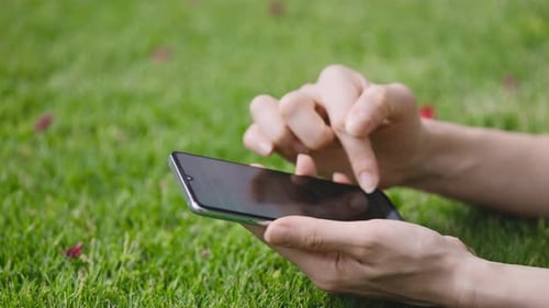 Women's Hands Use a Smartphone Shot Closeup A Woman is Lying on a Green Lawn and is Typing a Text or