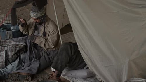 Un vagabond barbu répare un trou dans un pantalon, un sans-abri inconnu allongé dans une tente
