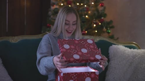 A woman wearing a cozy gray sweater opens a red gift box while sitting on a vintage green sofa. The
