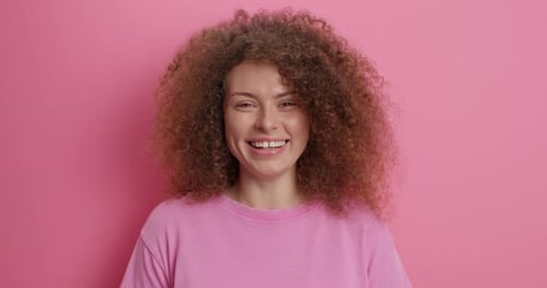 Smiling woman with curly hair poses on pink background
