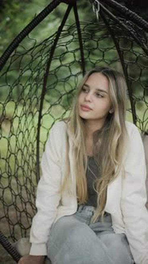 Young Woman with Long Blonde Hair Sitting in a Hanging Chair Outdoors