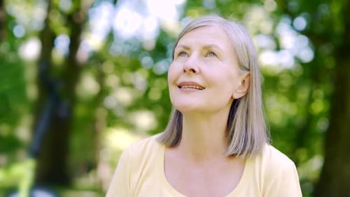 Close up portrait senior retiree woman standing in nature between forest trees relaxes, breathes