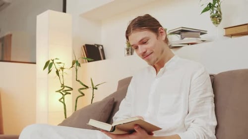 Young Man Dressed in Casual White Attire Sits Comfortably on a Couch Absorbed in Reading a Book