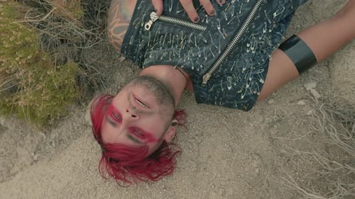Close up shot of a man with red hair and red eye makeup lying on a rocky desert ground