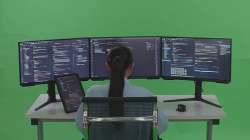 Rückansicht einer asiatischen Frau, die mit einem Tablet arbeitet, während sie Code mit mehreren Bildschirmen auf einem grünen Bildschirm schreibt