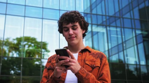 Happy Caucasian Man Uses a Smartphone While Standing on a City Street in Casual Clothes Background