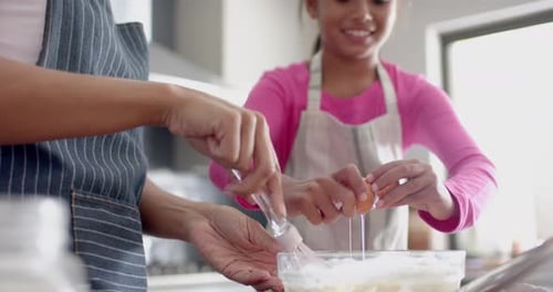 Happy biracial mother and daughter making cake mix, baking in kitchen, slow motion