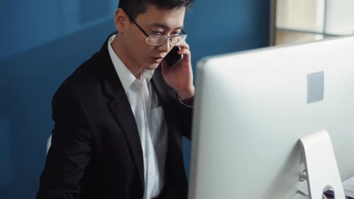 Retrato: Un joven chino con gafas usa la computadora para hablar por teléfono y escribir en el cuaderno de oficina