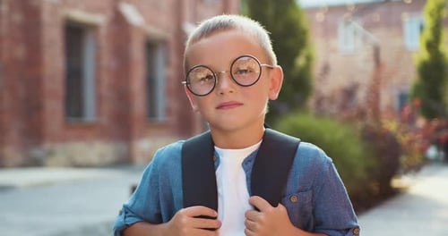Close-up portrait of little schoolboy in glasses and with backpacks standing on the school building