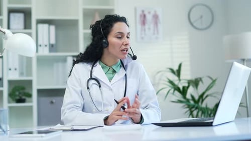 Confident female doctor in wireless headset talking on video call using laptop in modern hospital cl