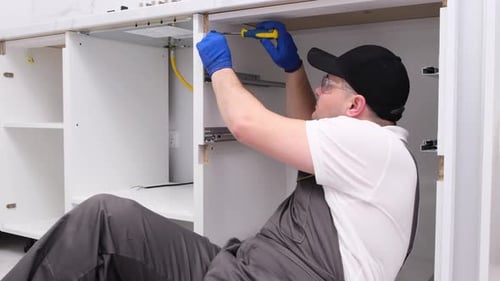 A Qualified Craftsman in Special Clothes Installs the Doors of a White Cabinet in a Modern Kitchen