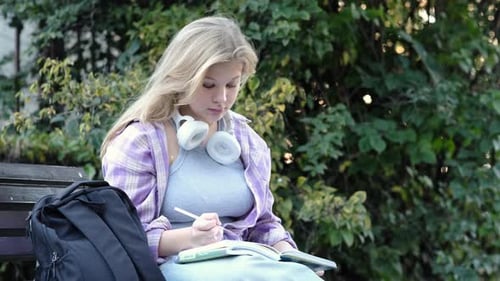 A Student Outdoors in a Park Immersed in Writing with a Backpack By Her Side and Headphones Around