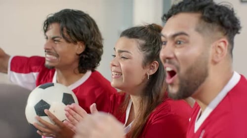 Game intensifying Diverse friends in jerseys gripping ball clapping popcorn cheering in living room