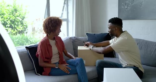 Couple afro-américain regardant un homme trancher du ruban adhésif et ouvrant une boîte sur un canapé, cadeau révélateur
