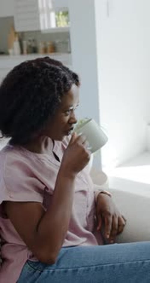 Steam rising from mug, woman seated on sofa inhaling, sipping tea, gazing toward window, relaxing