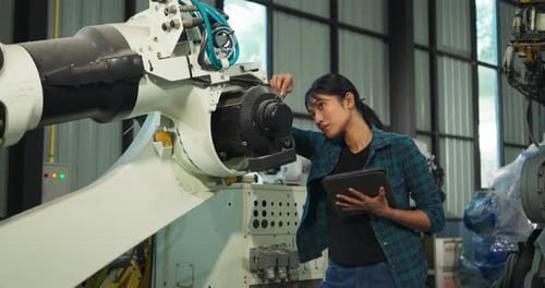 Woman Inspecting Robotic Arm with Tablet in Factory