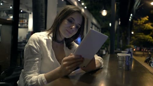 Woman Using Tablet Computer Sitting In Cafe At Night